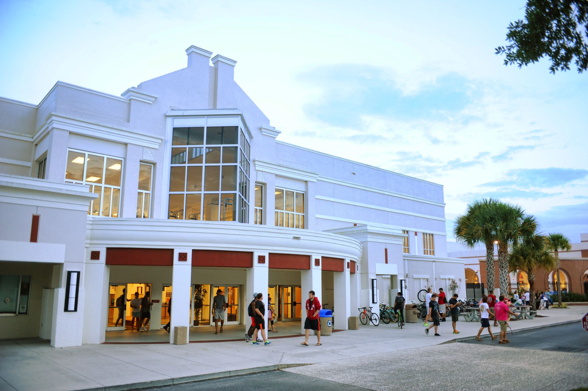 The exterior of the Clemente Center at Florida Tech during dusk, with students walking and biking in front of the building. The center features a prominent glass window section and palm trees nearby